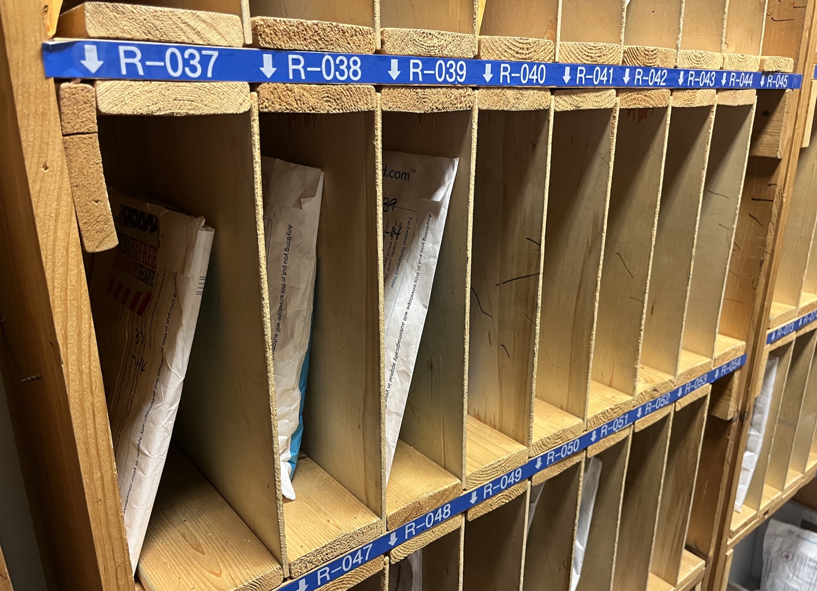 Mailroom-style sorting cubbies labeled R-037 through R-049, holding Magic Envelopes from individual customers awaiting processing